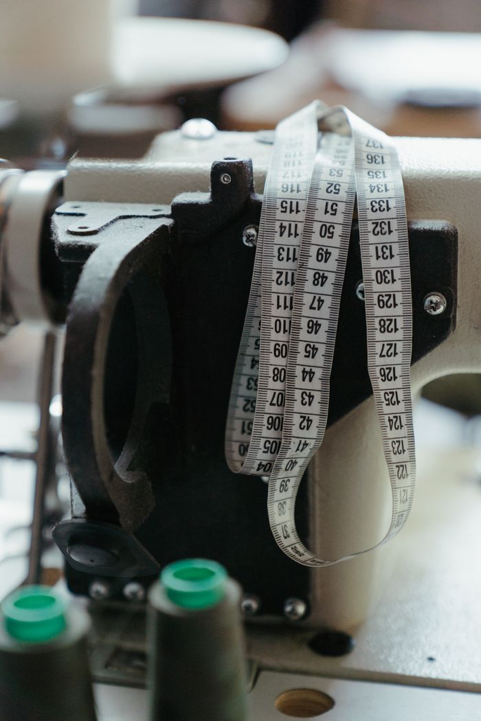 who-we-are Close-up of a sewing machine with measuring tape in a workshop, highlighting tailoring tools.