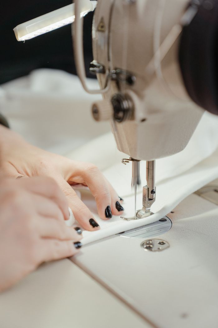 gallery-04 Detailed view of a sewing machine stitching fabric with visible hands, nails painted black.